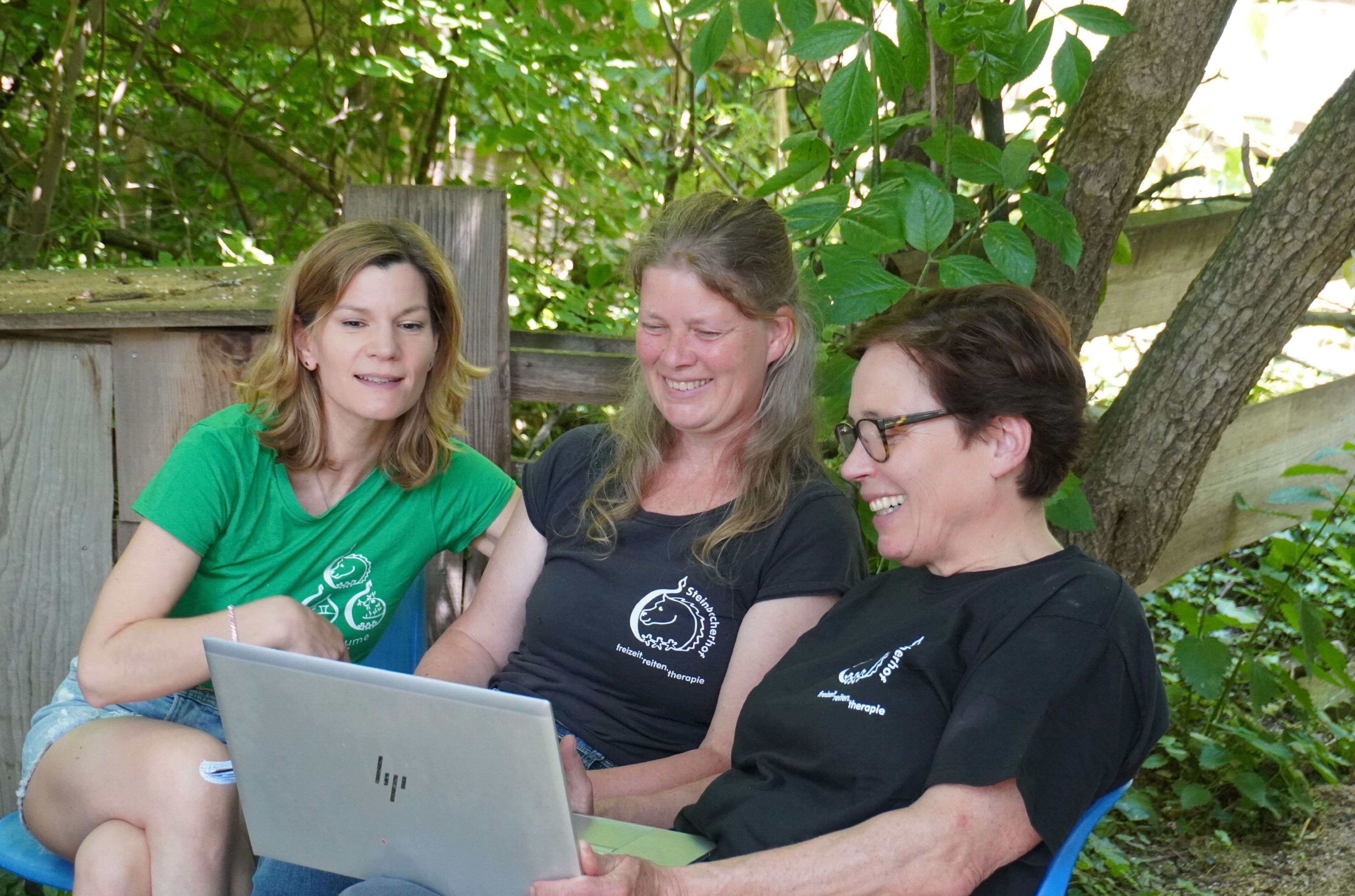 Drei Frauen sitzen im Freien in der Nähe eines Baumes, eine tippt auf einem Laptop. Sie tragen passende schwarze T-Shirts und scheinen in ein Gespräch vertieft zu sein. Grünes Laub und ein Holzzaun sind im Hintergrund zu sehen.