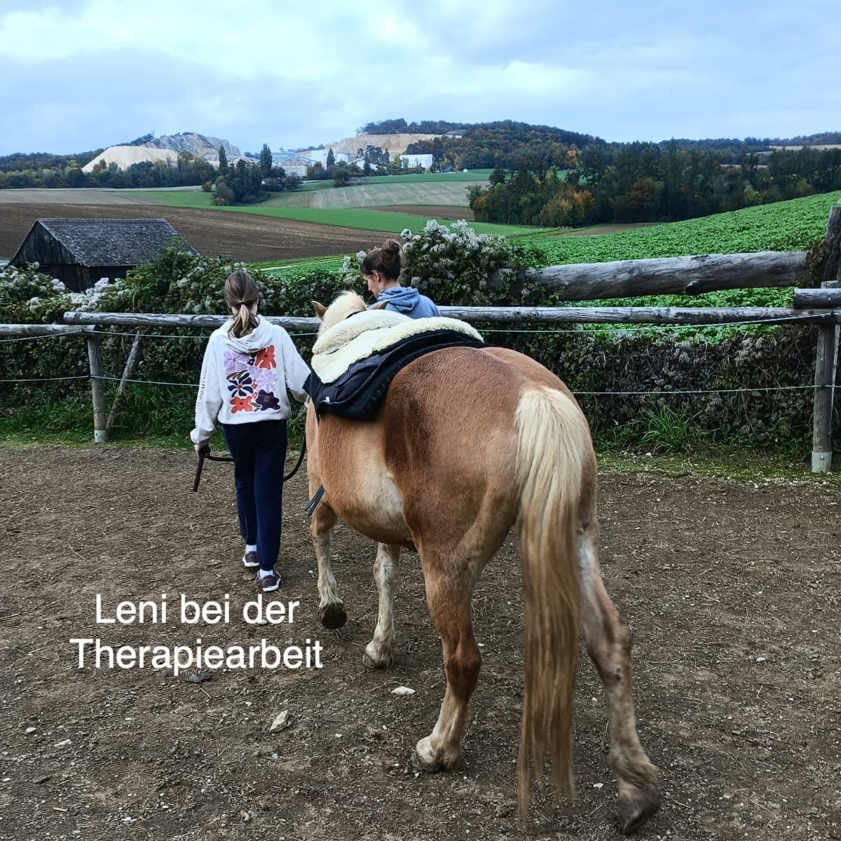 Zwei Frauen führen ein Pferd in einem eingezäunten Bereich, eine führt es, während die andere auf seinem Rücken reitet. Das Bild enthält den Text 'Leni bei der Therapiearbeit'. Der Hintergrund zeigt ein weites Feld mit Bäumen und einer entfernten Scheune.
