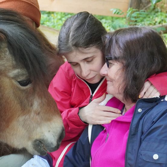 Ein junges Mädchen und eine erwachsene Frau, beide in Jacken gekleidet, trösten ein Pferd mit dunklen Augen, das in einem krautbewachsenen Bereich sitzt.