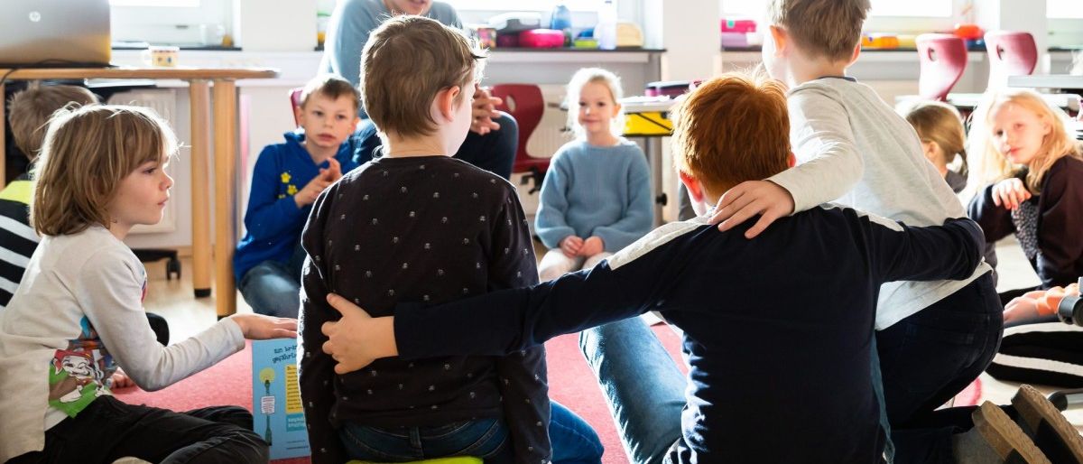 In einem Klassenzimmer sitzen mehrere junge Kinder im Kreis auf dem Boden und lauschen einem Mann, der vor ihnen steht. Ein Junge in einem blauen Shirt hält ein Buch. Hinter ihnen sitzt ein kleines Mädchen auf einem Stuhl, und ein Tisch ist mit verschiedenen Gegenständen bestückt.
