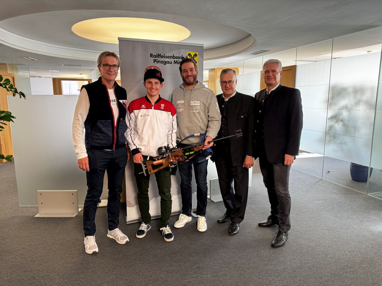 Fünf Männer stehen in einer modernen Büroumgebung zusammen. Einer hält eine Waffe. Hinter ihnen befindet sich ein Banner mit den Worten Raiffeisenbank Pinzgau Mit.
