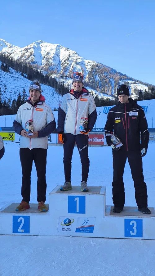 Drei Männer stehen auf einem verschneiten Podest und halten Trophäen. Sie tragen Winterkleidung und sind von verschneiten Bergen umgeben. Das Podest hat die Nummern 1, 2 und 3.