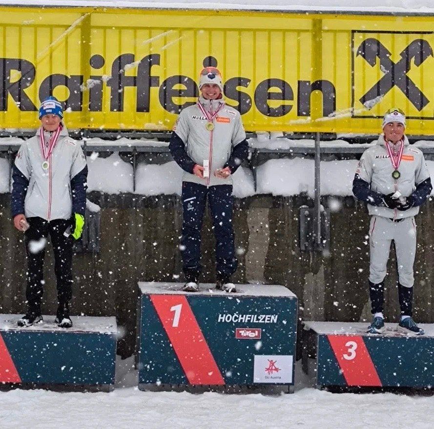 Drei Athleten stehen auf einem verschneiten Podest mit Medaillen um den Hals. Die mittlere Figur hält einen Pokal. Sie tragen Winterkleidung. Hinter ihnen steht ein gelbes Schild mit dem Text 'Raiffeisen' und einem Logo.