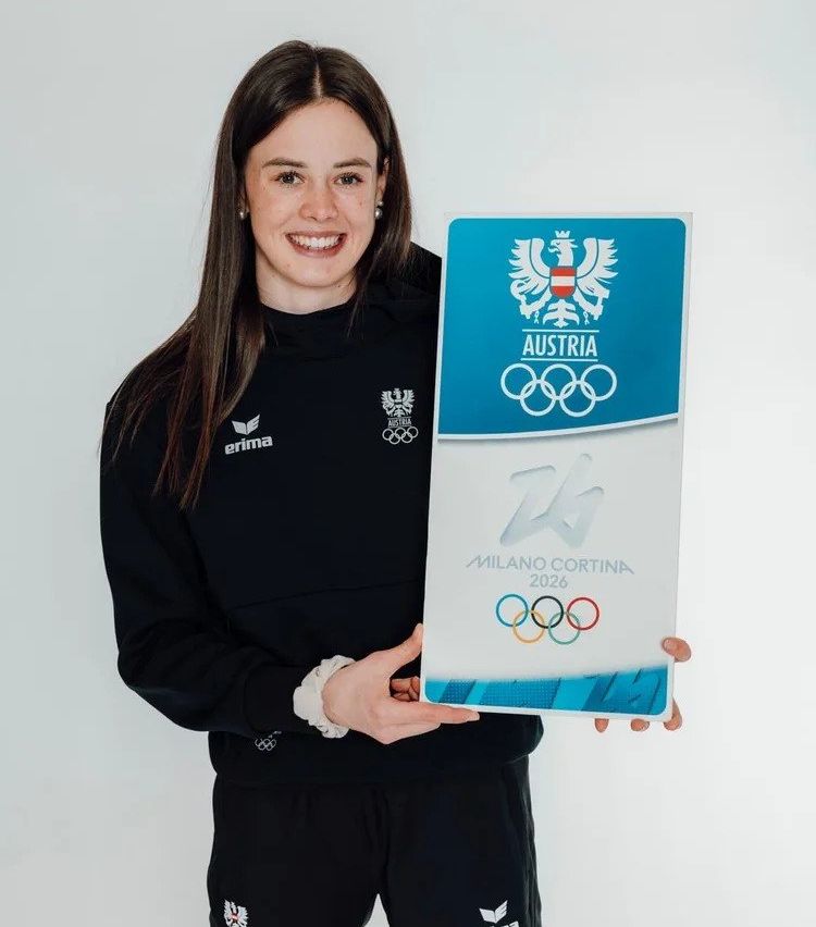 Eine Frau lächelt und hält ein Schild für die Olympischen Winterspiele 2026 in Mailand-Cortina, Italien, mit dem Olympischen Logo und der österreichischen Flagge.