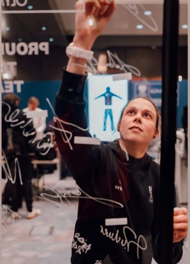 Eine Frau mit einem schwarzen Sweatshirt mit dem Wort 'eri' und einem Olympischen Logo schreibt auf eine Glaswand. Im Hintergrund befinden sich weitere Personen.