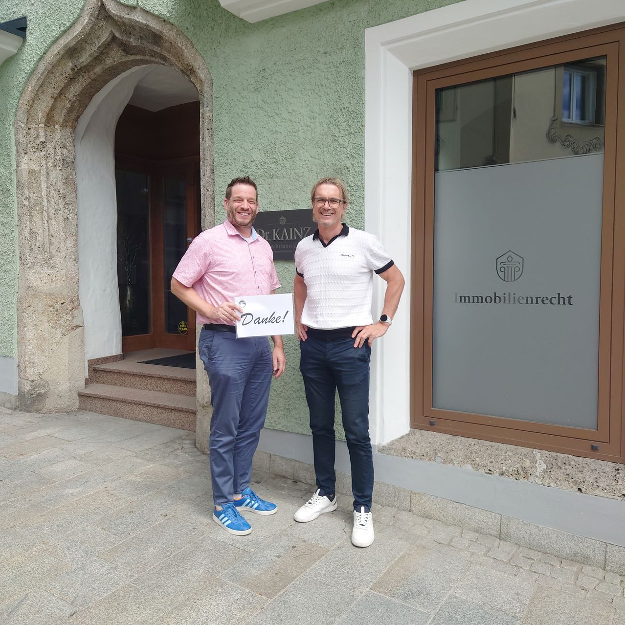 Zwei Männer stehen vor einem Gebäude mit einem Schild, das Danke! sagt. Ein Mann hält ein Schild, das Danke! sagt. Sie lächeln und scheinen für ein Foto zu posieren.