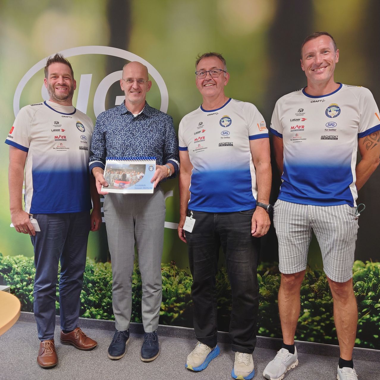 Vier Männer stehen zusammen vor einem grünen Hintergrund und lächeln für ein Foto. Der Mann in der Mitte hält ein gerahmtes Bild. Sie tragen alle weiße und blaue Trikots.