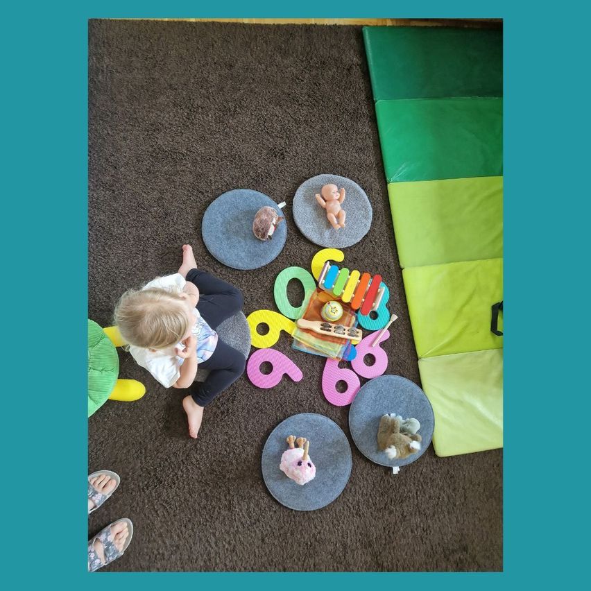Ein Kleinkind sitzt auf einem Teppichboden und spielt mit Spielzeug, umgeben von bunten Matten und einer Musikbox. Das Kind ist barfuß und scheint die Spielzeuge zu genießen.