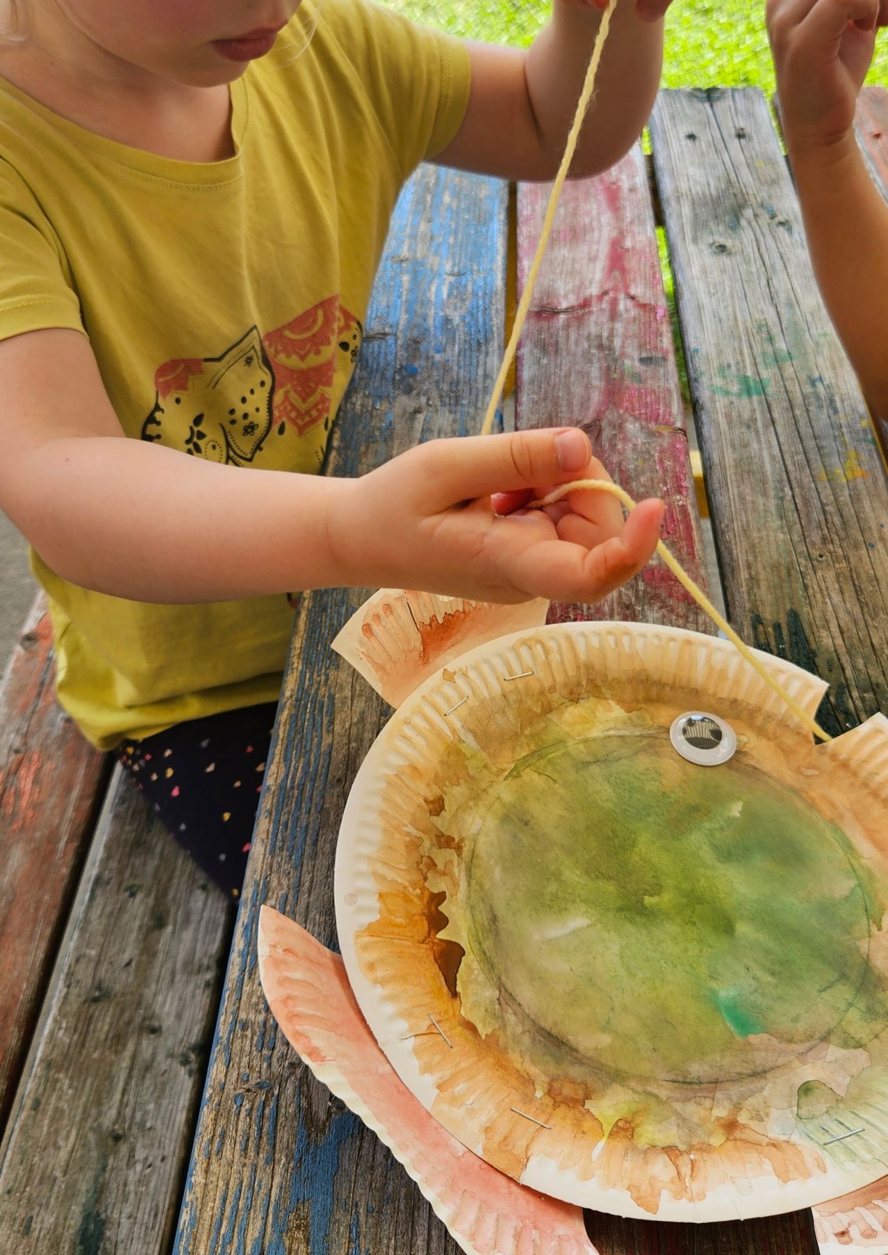 Ein Kind sitzt auf einer Picknickbank und hält eine Schnur, die an einen Papiertellerfisch befestigt ist. Der Fisch ist mit Wasserfarben in Grün- und Orangetönen bemalt.