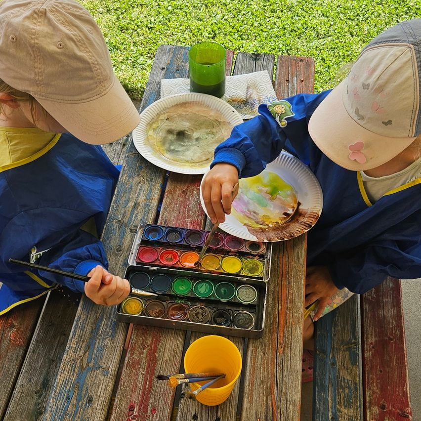 Zwei Kinder in Mützen malen auf Tellern an einem Picknicktisch. Ein Farbpalette mit verschiedenen Farben steht vor ihnen offen.