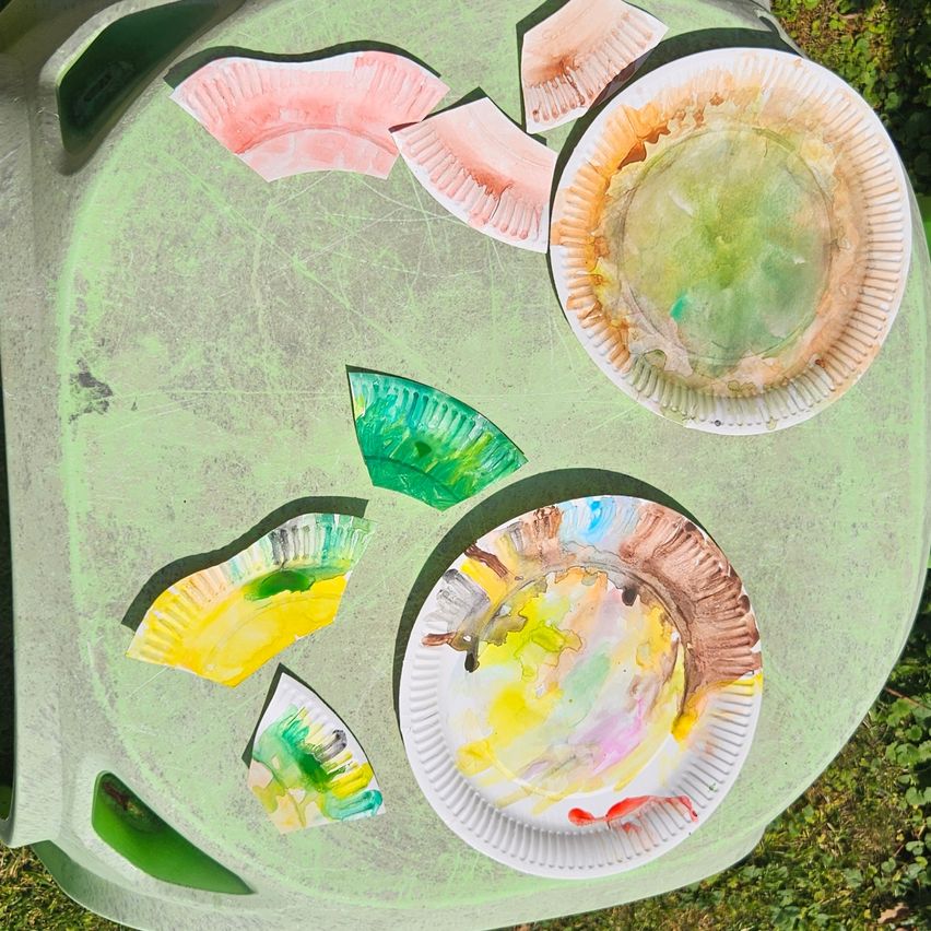 Mehrere Pappteller mit unterschiedlichen Farben und Mustern, einige zerbrochen, liegen auf einem grünen Tablett draußen auf einer Grasfläche.