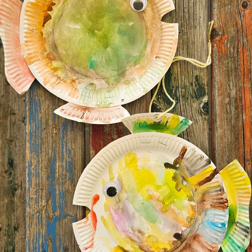 Zwei Papiertellerfische, mit verschiedenen Farben bemalt, hängen vor einem hölzernen Hintergrund. Die Fische haben Augen und Flossen, aus Papier gefertigt.