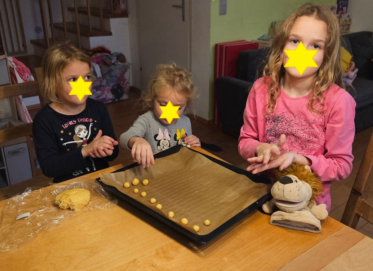 Three girls are sitting around a table, each wearing a yellow star on their faces. The middle girl is arranging dough balls on a baking sheet. A stuffed lion is nearby.