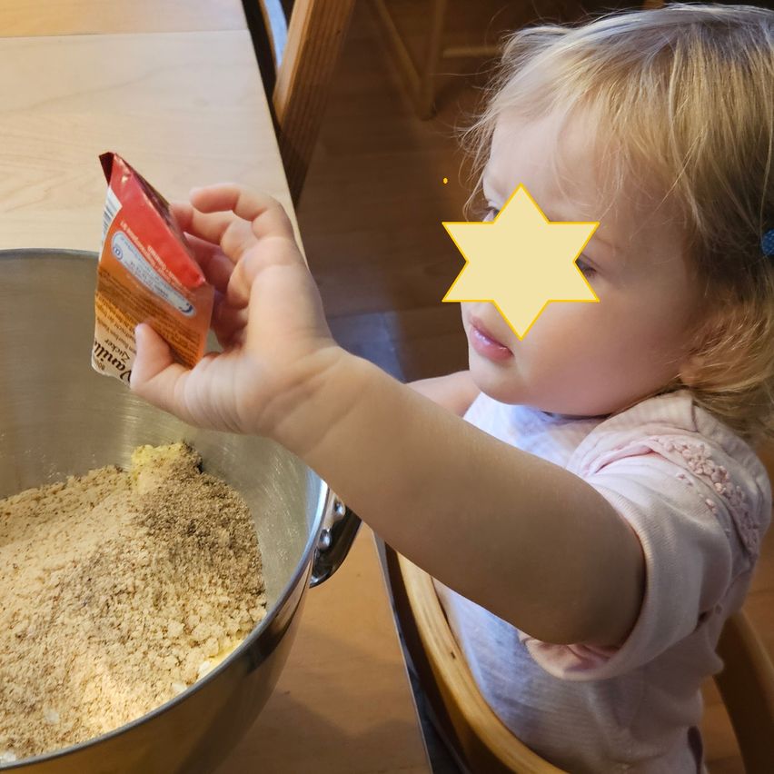 A young girl is adding dry ingredients to a bowl on a wooden table. She is holding a packet of oats.