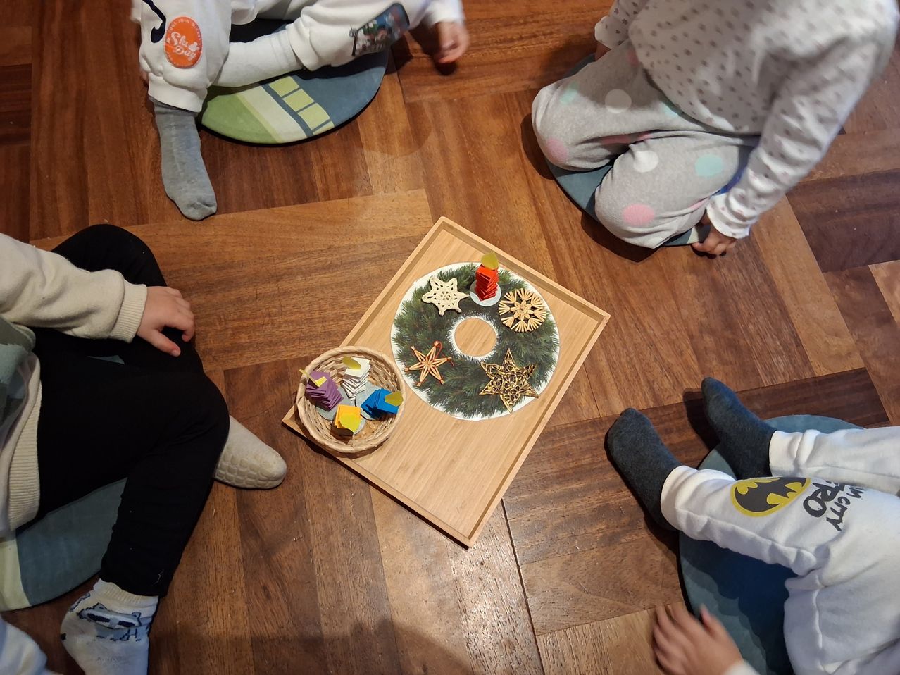 Several children sit in a circle on the floor. A wooden board has a Christmas wreath and ornaments on it. A basket of colorful cubes is next to the board.
