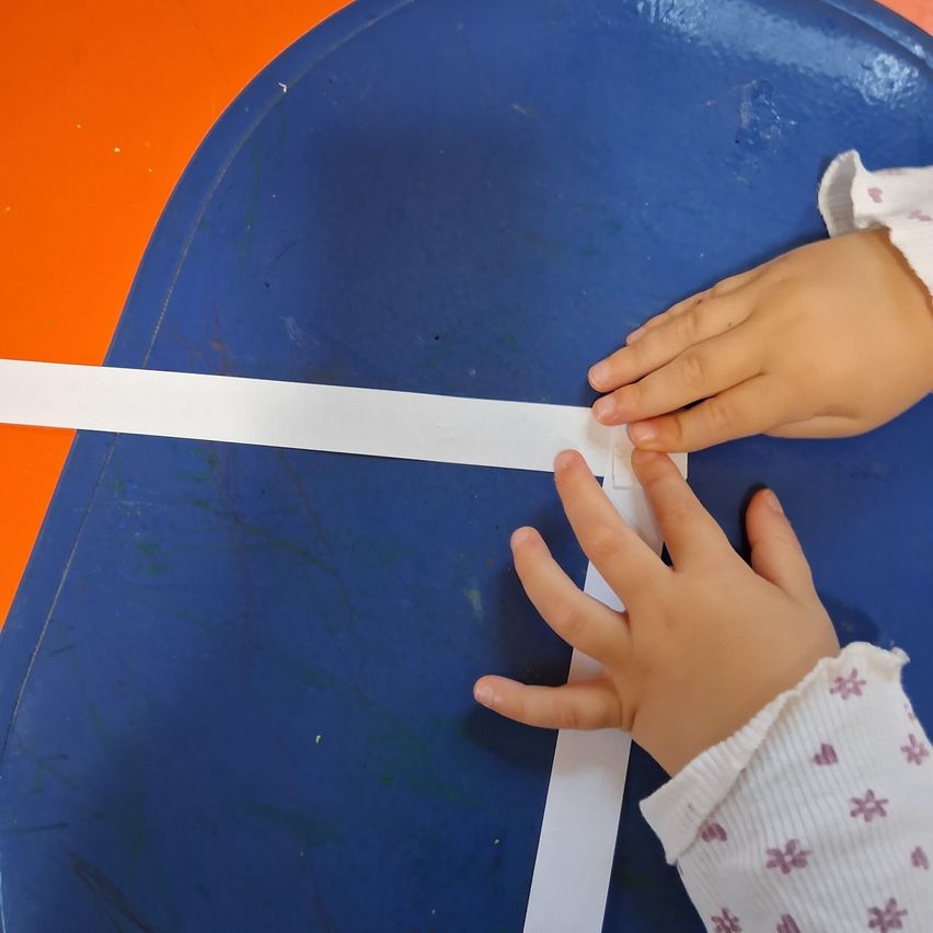 A child is folding a white paper strip on a blue tray, possibly for an art activity. The child's hands are visible, and the background shows a mix of orange and blue.