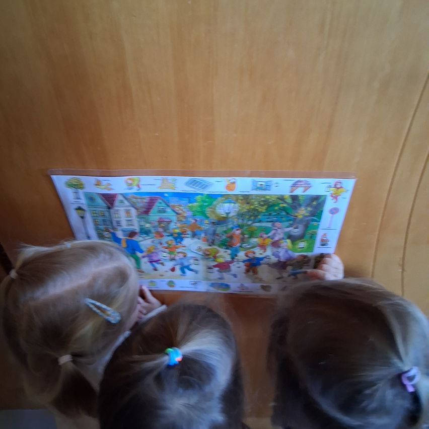 Three young girls with ponytails are looking at a puzzle on a wooden wall. The puzzle shows a colorful scene with buildings, trees, and people.