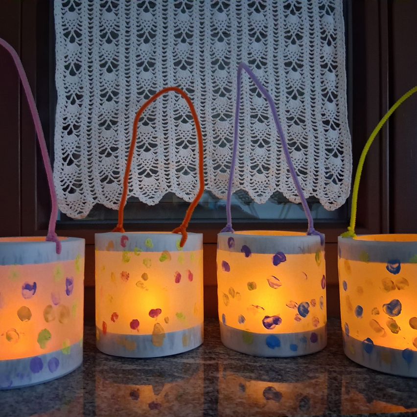 Four homemade lanterns with handles are lined up on a countertop. They are painted with colorful dots and contain candles that are lit.