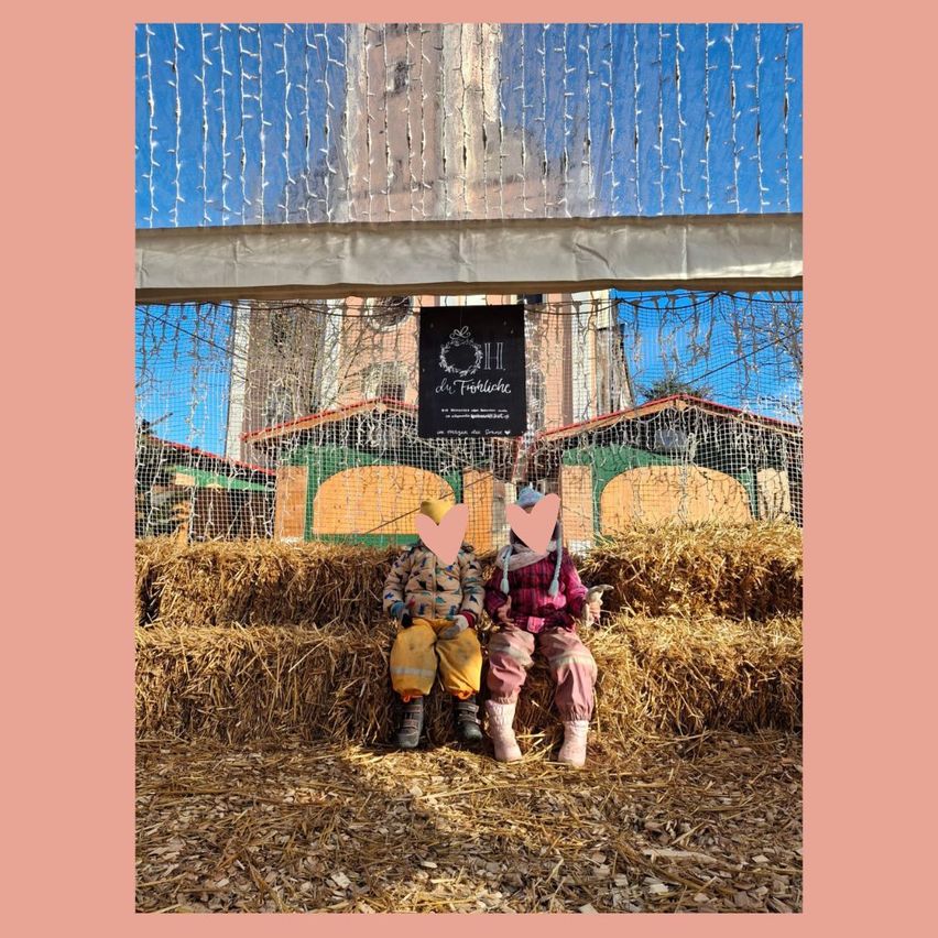 Zwei Kinder sitzen auf einem Heuballen mit roten Herzen auf den Köpfen, vor einem dekorativen Schild und umgeben von herbstlichen Dekorationen.