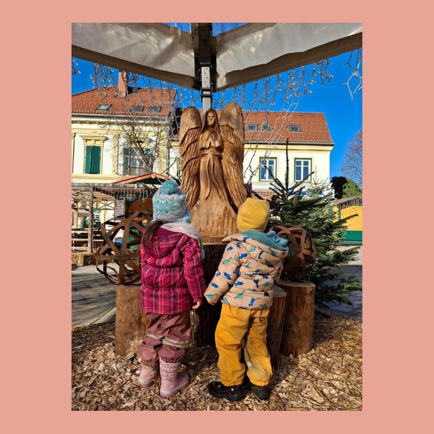 Zwei Kinder stehen vor einer hölzernen Engelsstatue unter einem Baldachin, mit Weihnachtsbäumen und einem Haus im Hintergrund.