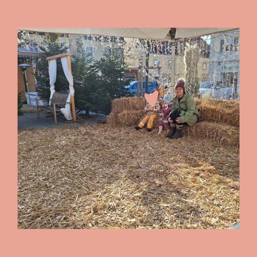 Eine Frau mit zwei Kindern sitzt auf Heuballen, mit einem Weihnachtsbaum und einem Pavillon im Hintergrund.