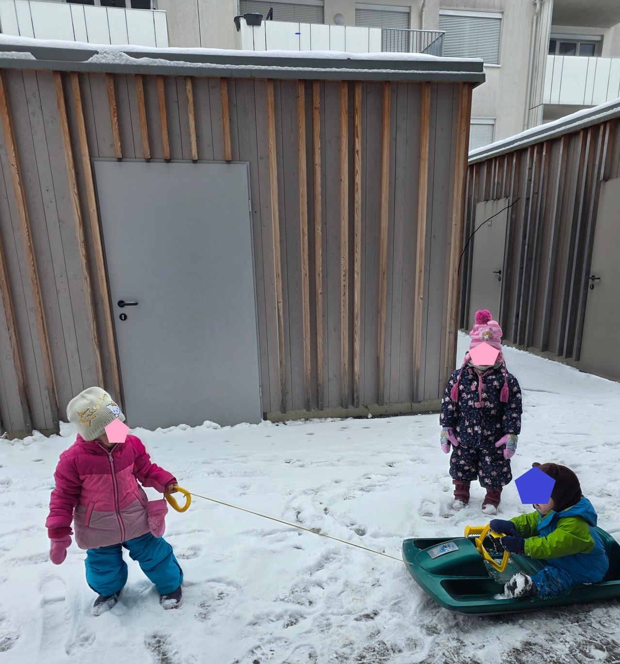 Drei Kinder sind draußen im Schnee, zwei ziehen einen Schlitten, auf dem ein drittes Kind sitzt. Sie sind in Winterkleidung und Mützen gekleidet. Hinter ihnen steht ein Gebäude mit einer weißen Tür.