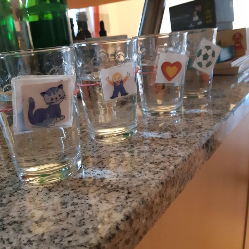 Vier klare Gläser mit Wasser stehen auf einer Granitarbeitsplatte. Jedes Glas hat einen Aufkleber mit verschiedenen Bildern: eine Katze, ein Junge, ein Herz und einen Fußball.