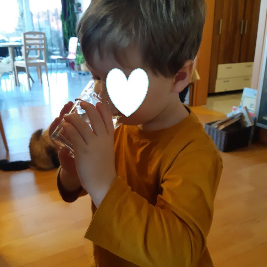 Ein kleiner Junge trinkt Wasser aus einem Glas. Ein Kater sitzt hinter ihm. Ein herzförmiger Aufkleber befindet sich im Gesicht des Jungen.