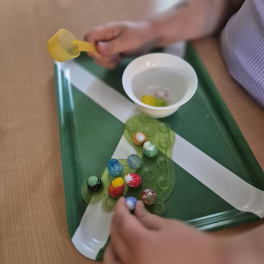Eine Person spielt mit bunten Murmeln auf einem grünen Tablett und benutzt einen gelben Löffel, um sie zu bewegen.