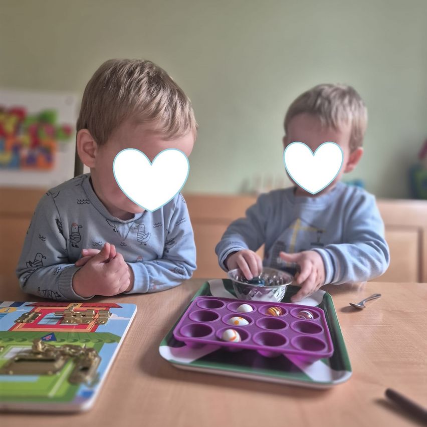 Zwei Jungen sitzen an einem Tisch, einer spielt mit Spielzeug, während der andere eine Schüssel mit Murmeln hat.