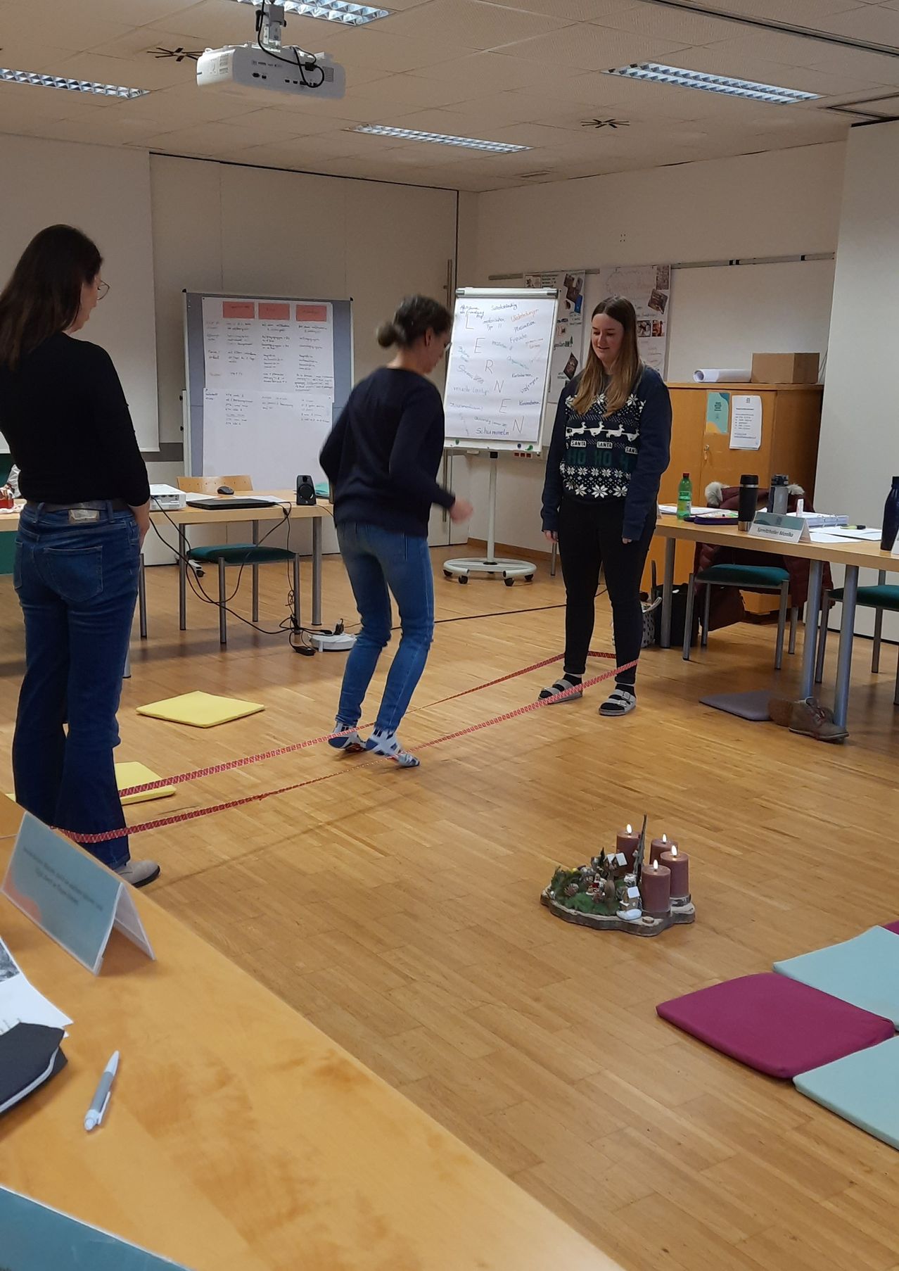 Three women in a room, with one holding a red rope. A whiteboard and various items on a desk. The floor has a candle display.