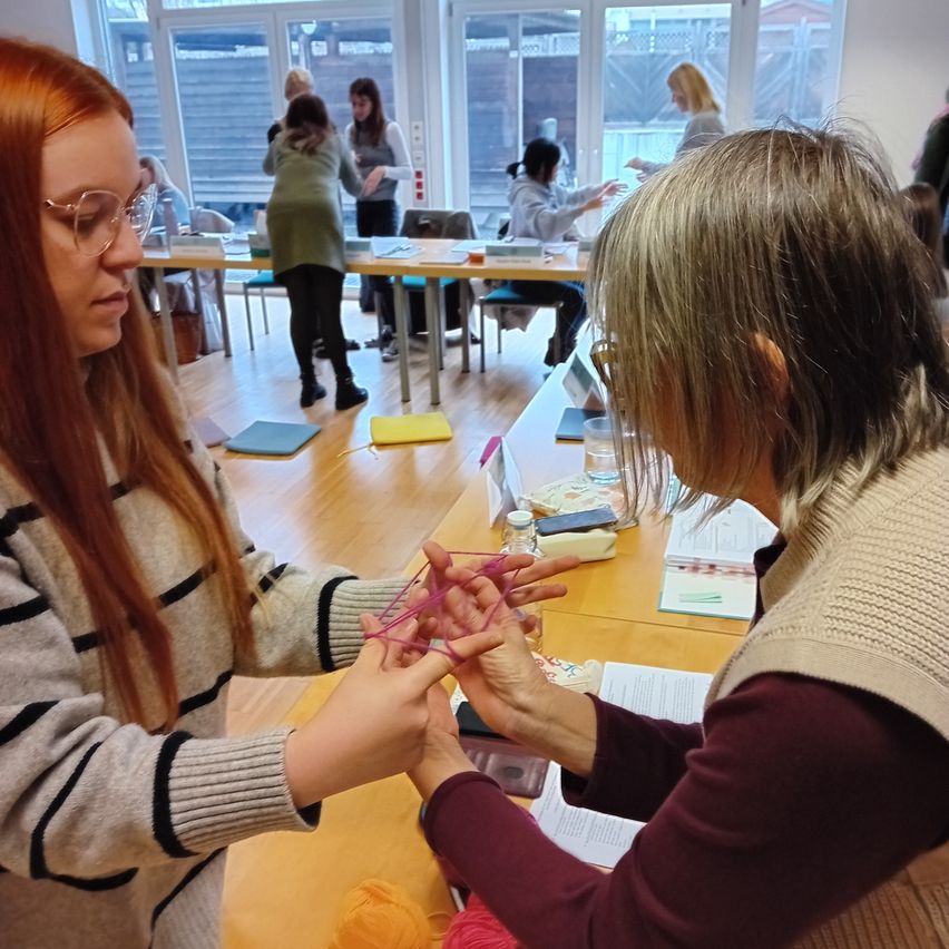 In a classroom, a woman with glasses and long hair works with pink yarn on a project, while an older woman assists. Other students are present, engaged in various activities.