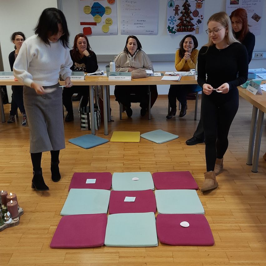 In a classroom, several women are seated around a table with various items on it. Two women are standing in front of the table, one holding a small object. The room has a wooden floor and a white wall.