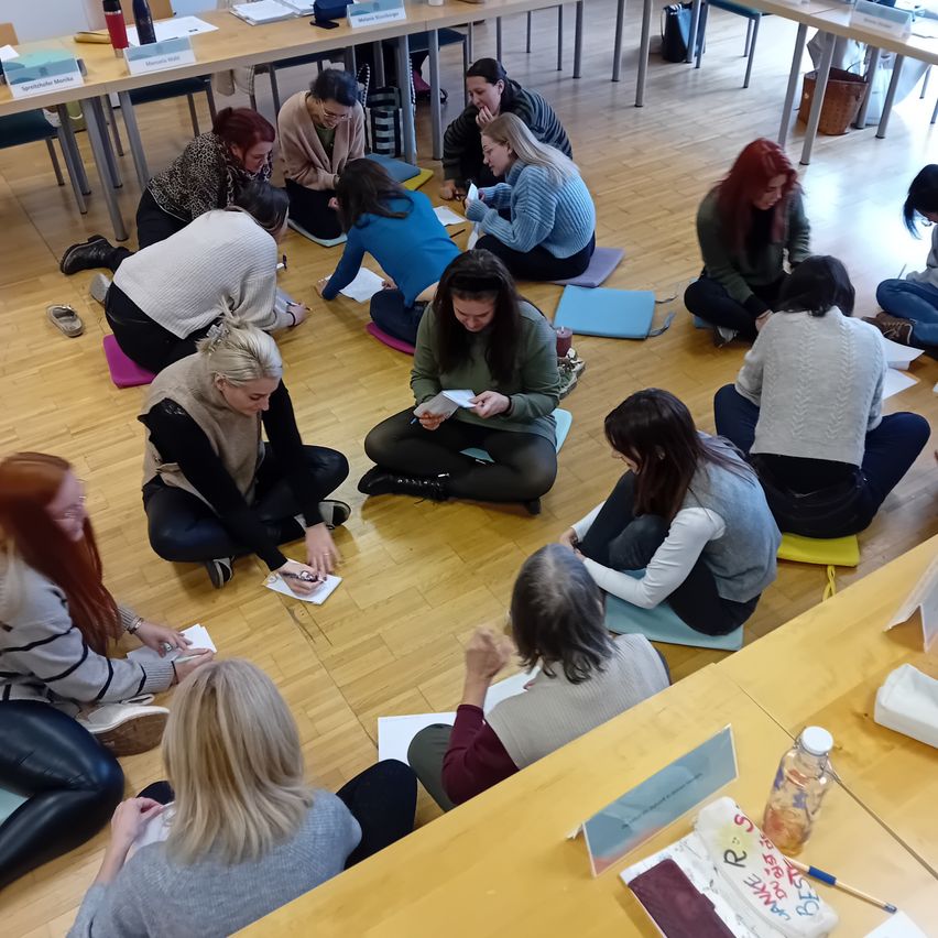 A group of women are sitting in a circle on the floor, engaged in an activity, possibly a workshop. They are holding papers and some are writing. There are tables with papers and pens.