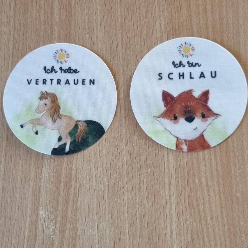 Zwei runde Aufkleber auf einer Holzoberfläche. Einer zeigt ein Pferd mit dem Text 'Ich habe Vertrauen'. Der andere zeigt einen Fuchs mit dem Text 'Ich bin SCHLAU'.