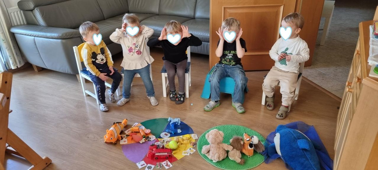 Drei junge Kinder mit Herzaufklebern auf dem Gesicht sitzen in Stühlen um Spielzeug auf dem Boden. Ein Kind in einem gelben Hemd sitzt neben ihnen.