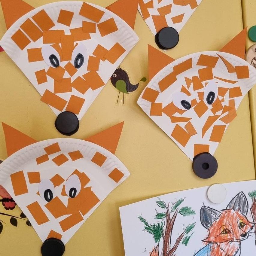 Vier Fuchsgesichter aus Papiertellern und Karton mit orangefarbenen Quadraten schmücken einen gelben Hintergrund. Jedes Gesicht hat Augen und eine Nase. Ein Bild von einem Fuchs und einem Baum befindet sich in der Nähe.