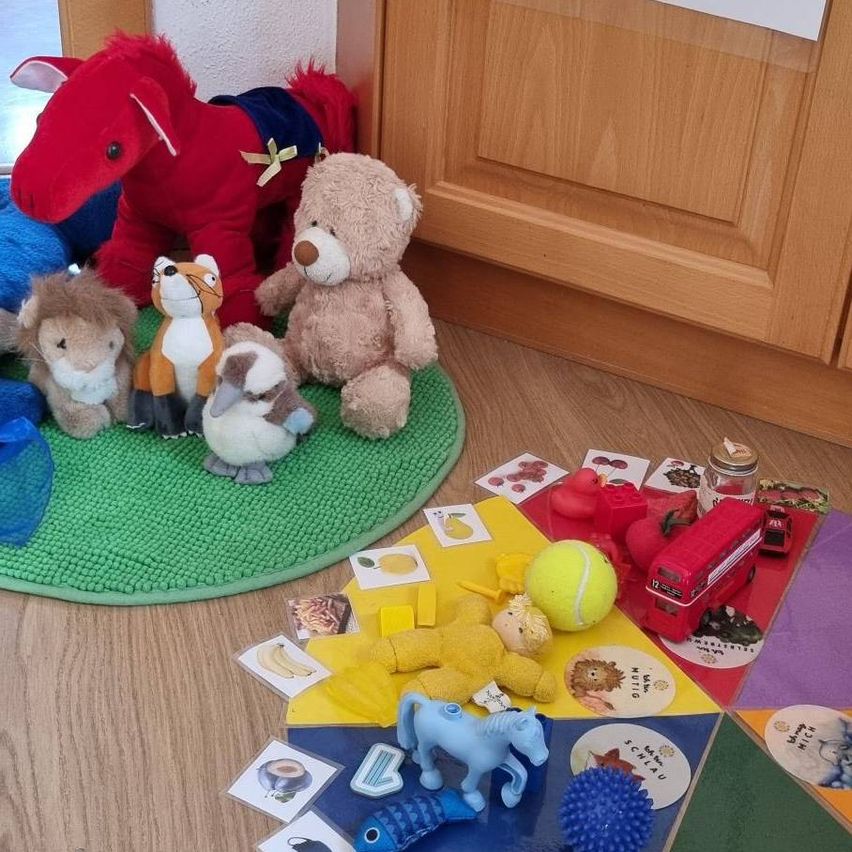Ein grüner Teppich hat Plüschtiere, darunter ein Fuchs, Löwe, Bär und Pferd. Ein mehrfarbiger Teppich mit Bus, Tennisball und anderen Spielzeugen daneben.