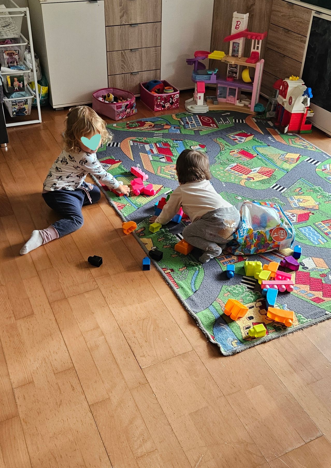 Two toddlers are playing on a colorful floor mat with toys. One sits, and the other stands, holding a toy. Various toys are scattered around.