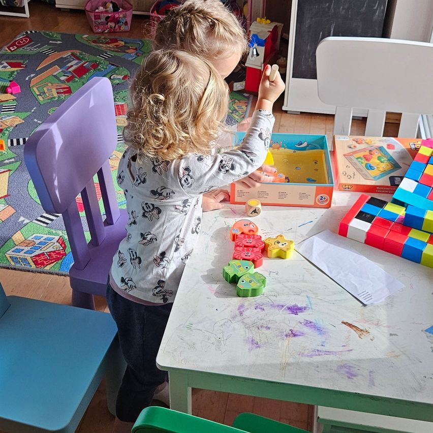 Two young girls are playing with toys on a table, one girl is reaching for a toy. The table is filled with colorful toys and books.