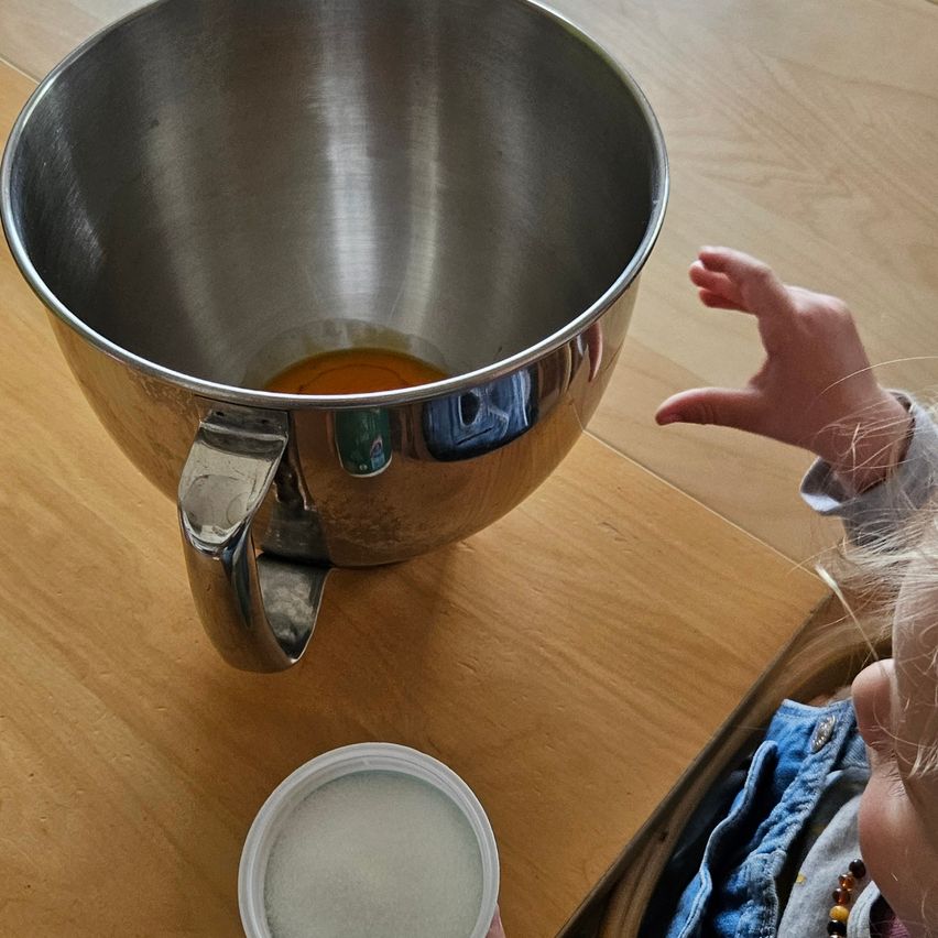 A child reaches towards a bowl of batter on a wooden table, a container of sugar beside it.
