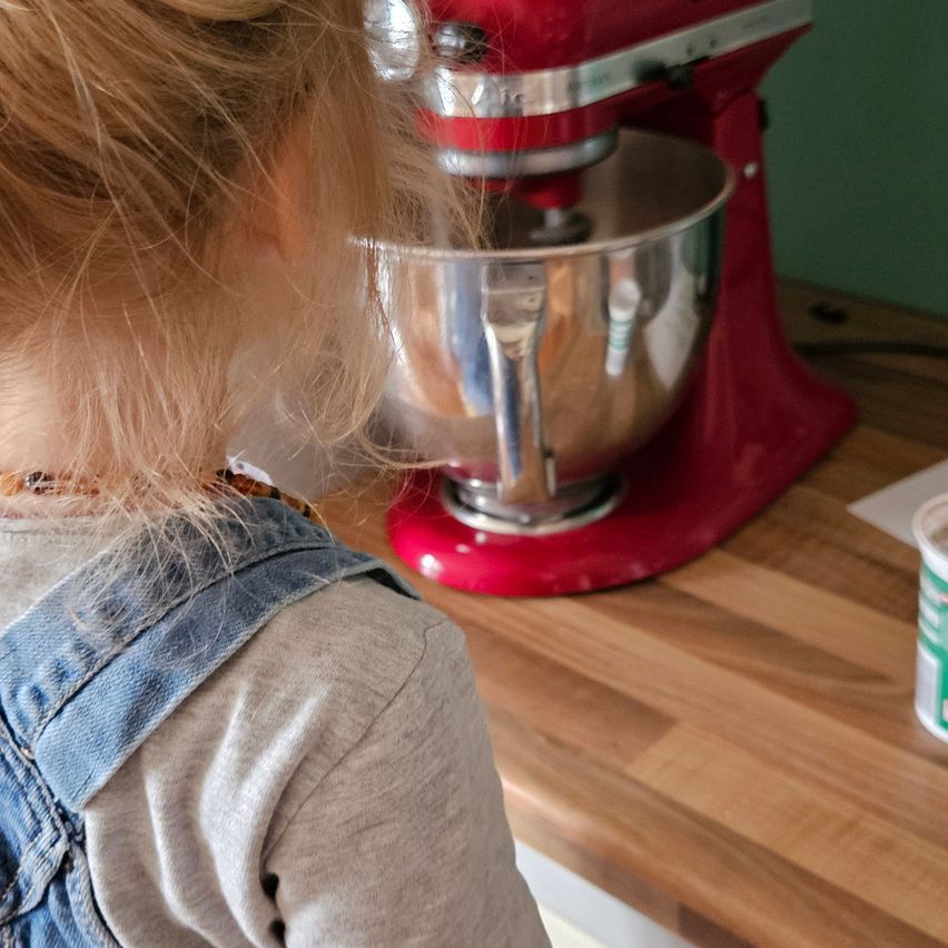A young girl wearing a denim jumper is standing in front of a red mixer on a wooden counter. A cup with a green lid is on the counter.