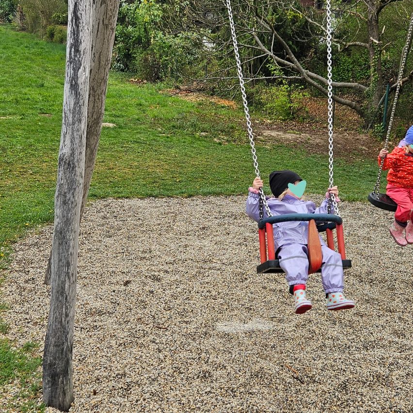 Two children are playing on swings in a park. One is wearing a mask, while the other is in a red outfit.