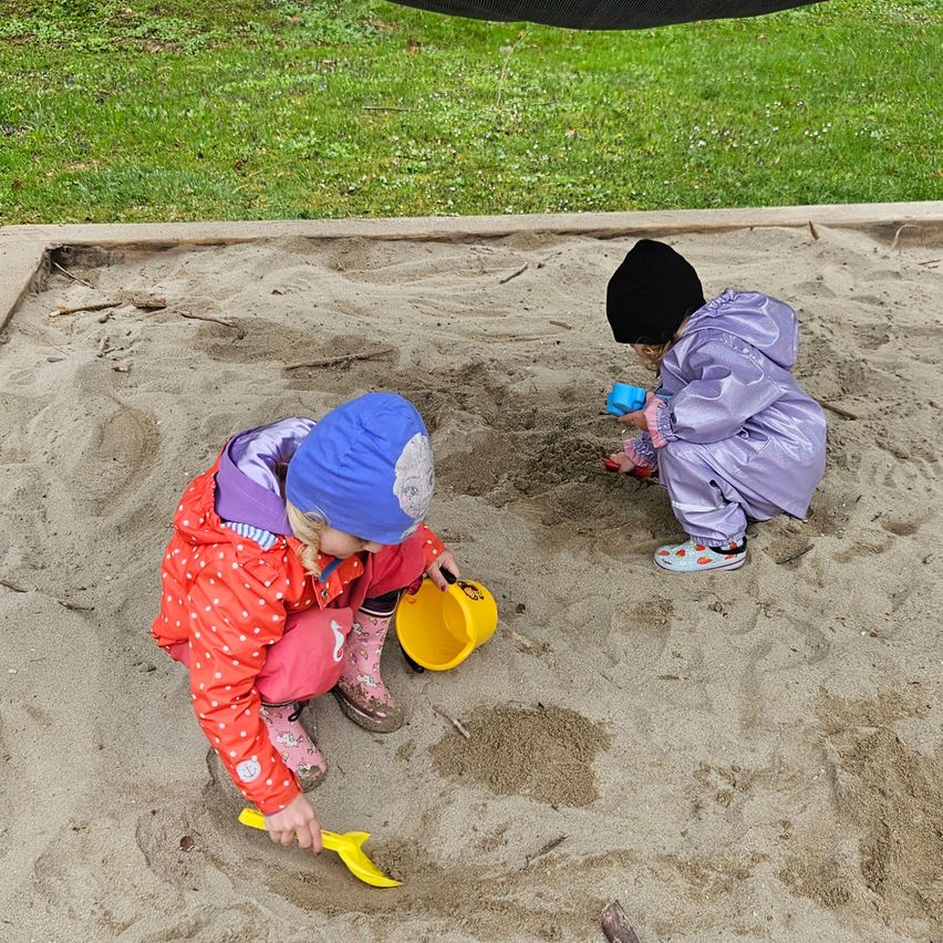 Two young girls are playing in a sandbox, one with a yellow bucket and the other with a blue toy. Both are wearing rain gear and boots.