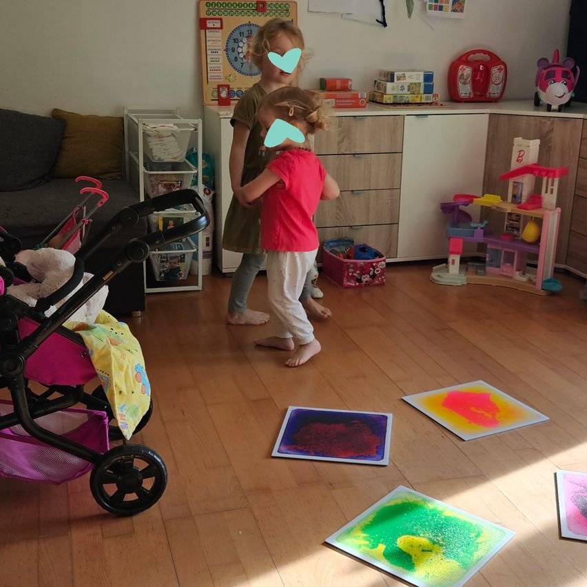 Two young girls play on a wooden floor with colorful paint on paper. A stroller, couch, and toys are in the background.
