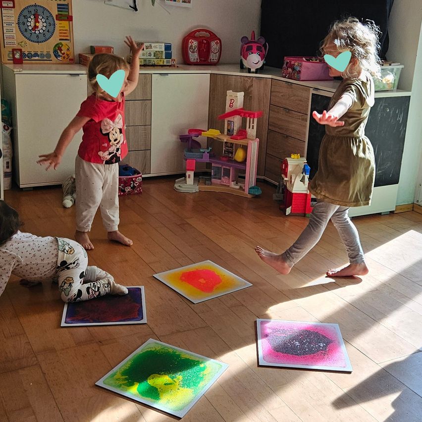 Three children are playing on the floor with four colorful paintings. One child is standing on tiptoes, another is kneeling, and the third is standing with her arms raised.