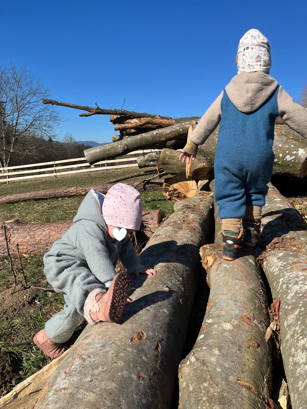 Zwei Kinder in Winterkleidung gehen auf Baumstämmen. Ein Kind trägt einen rosa Hut mit einem Herz darauf. Hinter ihnen befindet sich ein Holzstapel und ein Zaun. Der Himmel ist klar.