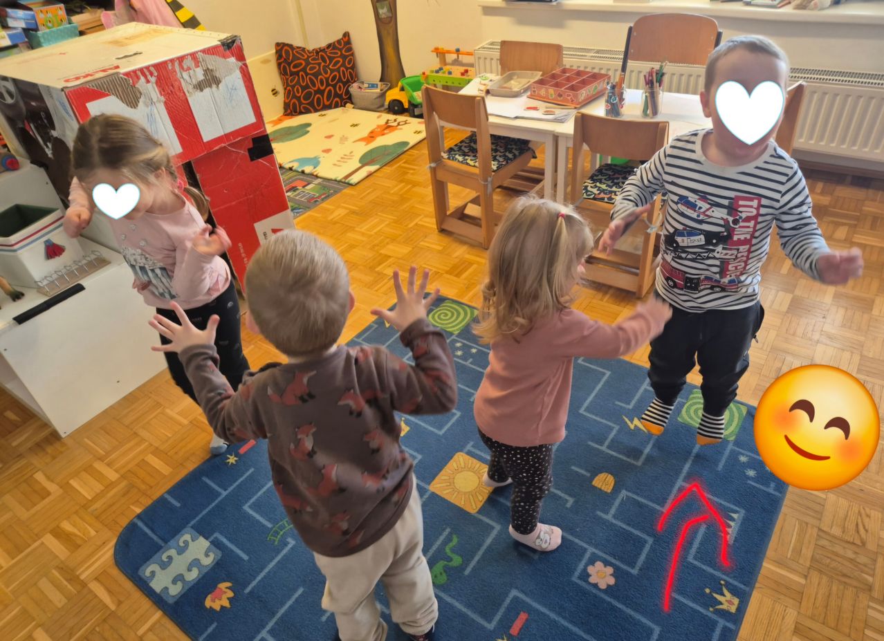 Four children play in a room with a blue carpet. One boy wears a fox-printed shirt. A girl stands with her arms outstretched. Another boy wears a striped shirt. The room has a wooden floor, chairs, and a table with toys.