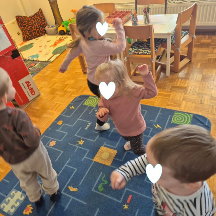 Four children are playing on a blue carpet with a maze design, surrounded by a room with wooden floors, chairs, and a table with art supplies. Two girls in pink long sleeves dance with hearts above their heads.