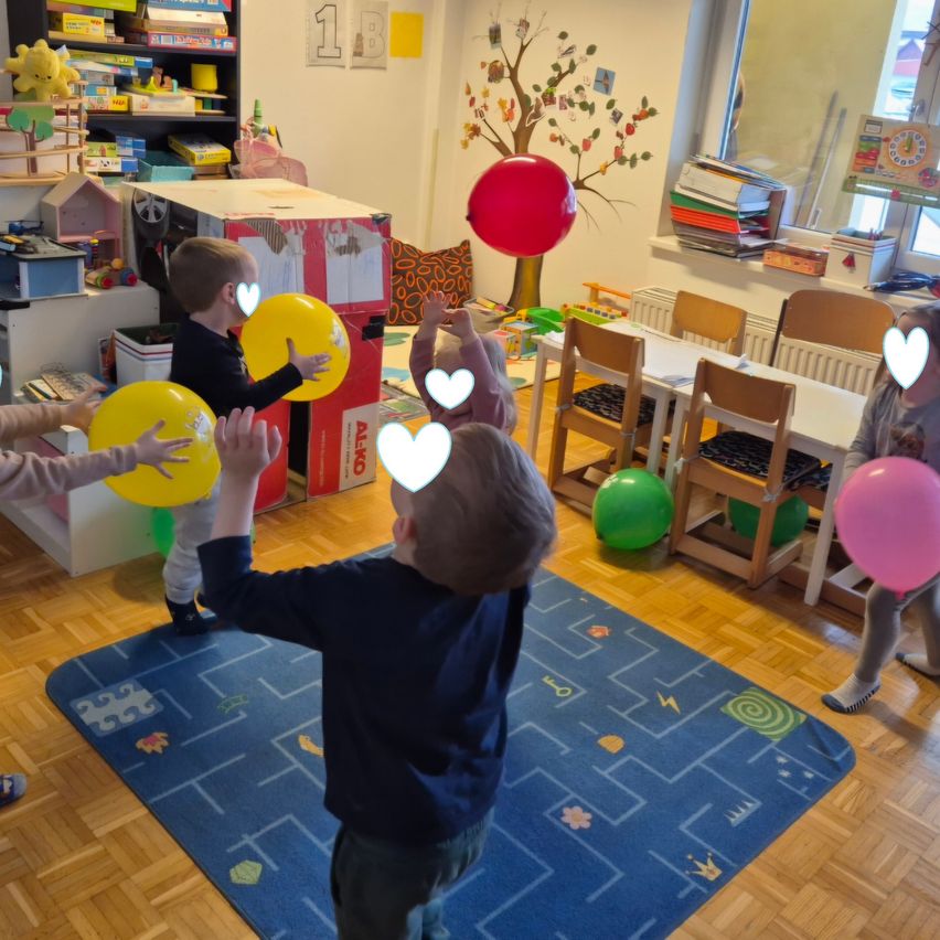 Several children play with balloons in a brightly lit room with wooden floors and various toys. One child is holding a red balloon.